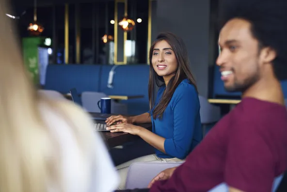 A woman smiles while working on a laptop in a modern cafe setting