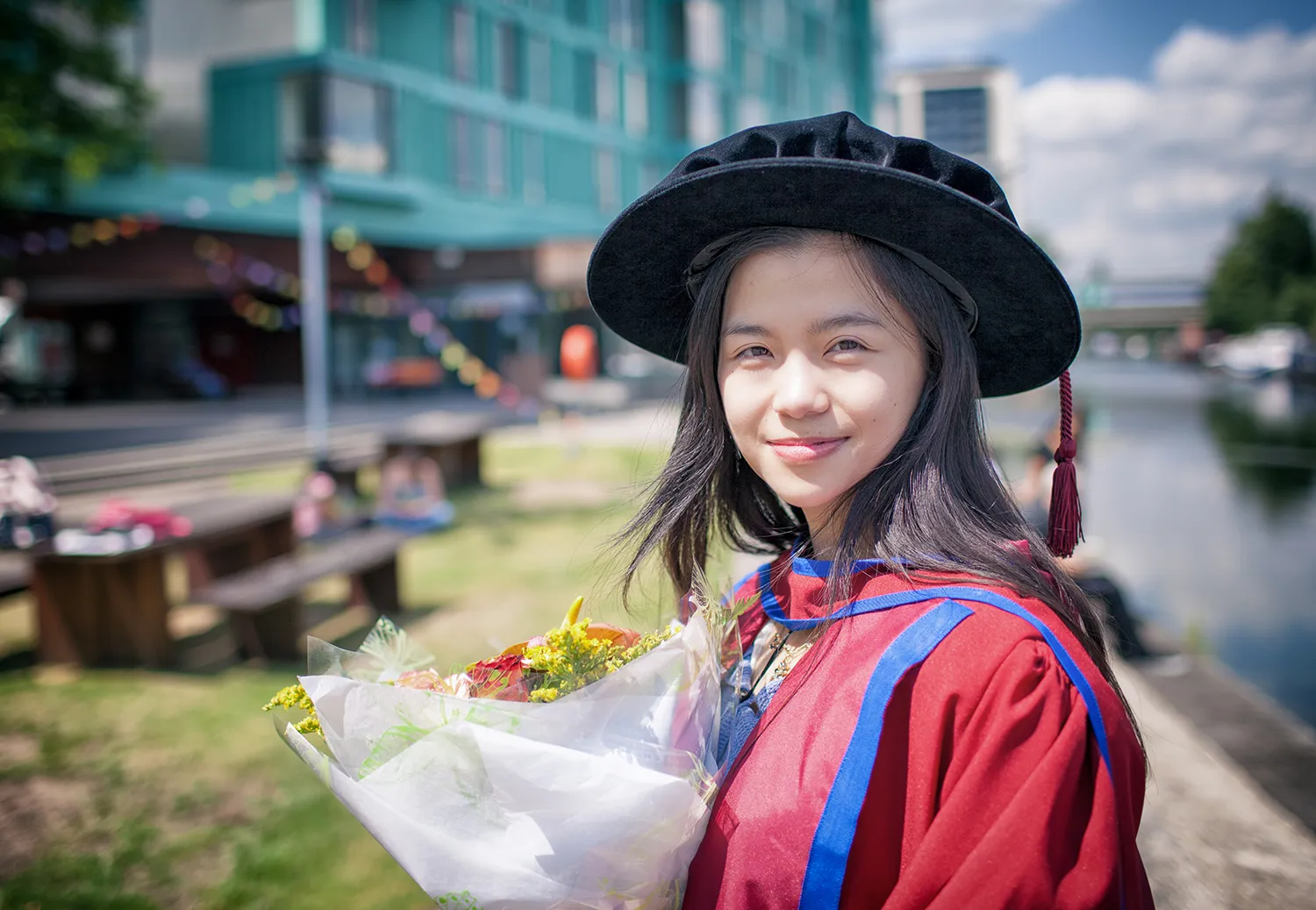 A graduate wearing academic dress and holding a bouquet beside a canal.
