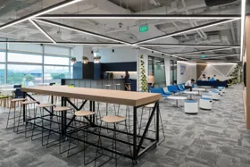 Modern office interior featuring a communal workspace with seating, a kitchen area, and large windows