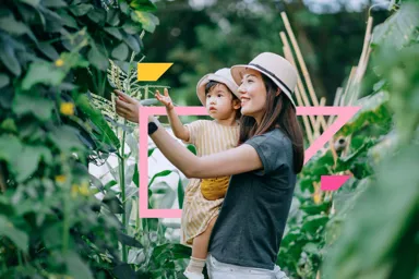 A woman holds a child in a garden surrounded by green plants