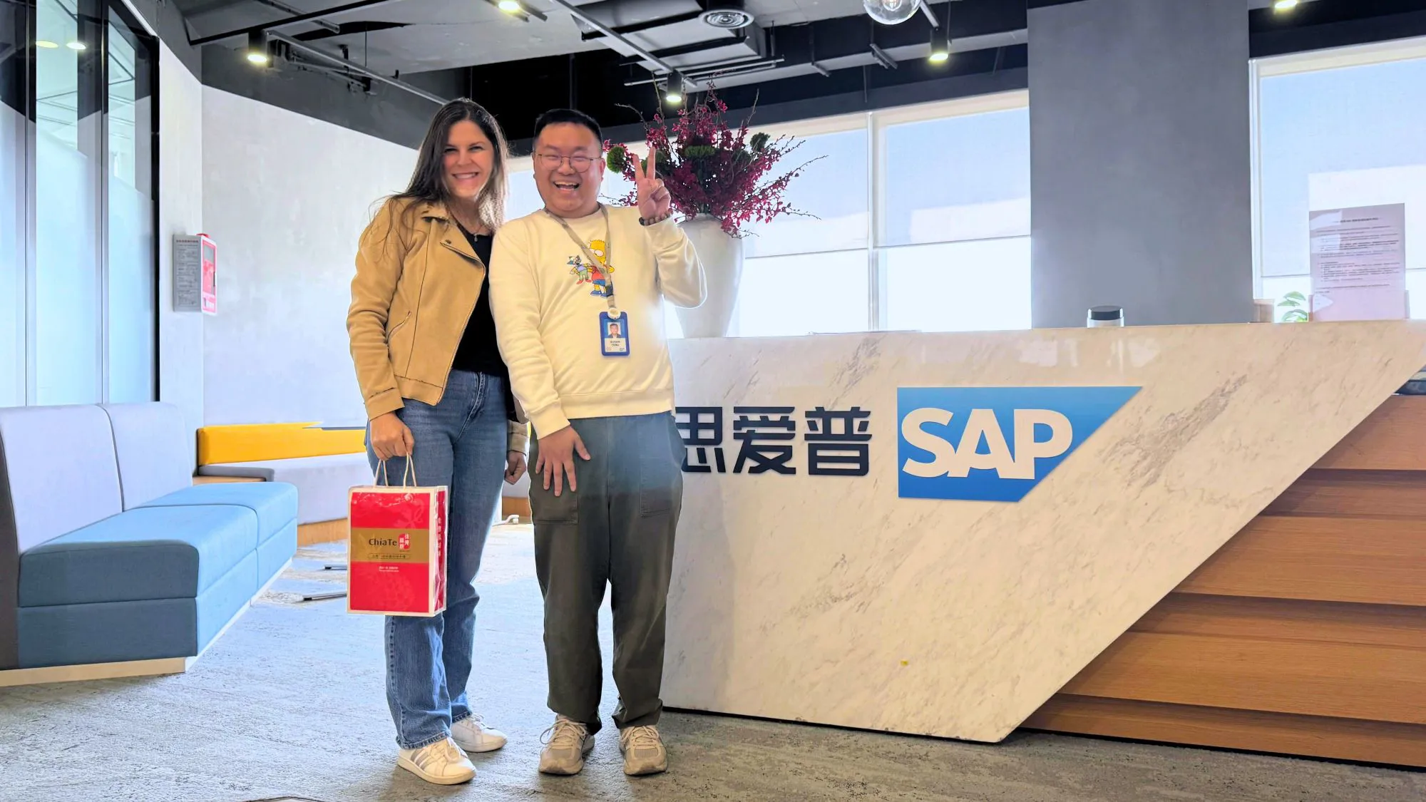 A woman wearing a tan jacket stands next to a man in a white sweater at a reception area with a large marble counter featuring the SAP logo. The background includes a gray sofa and decorative flowers.