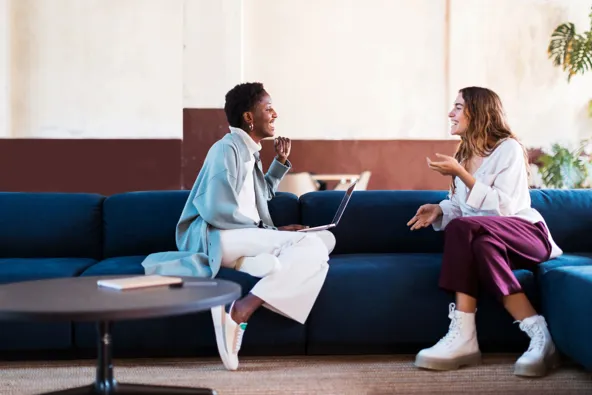 Two women sitting on a couch, engaged in conversation with a laptop