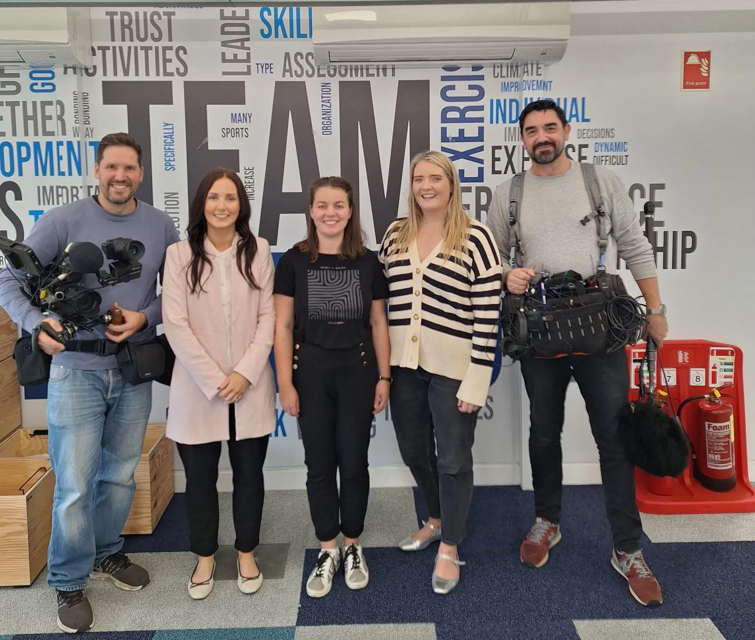 Five people standing together in front of a wall featuring teamwork-related words, with two holding video equipment.