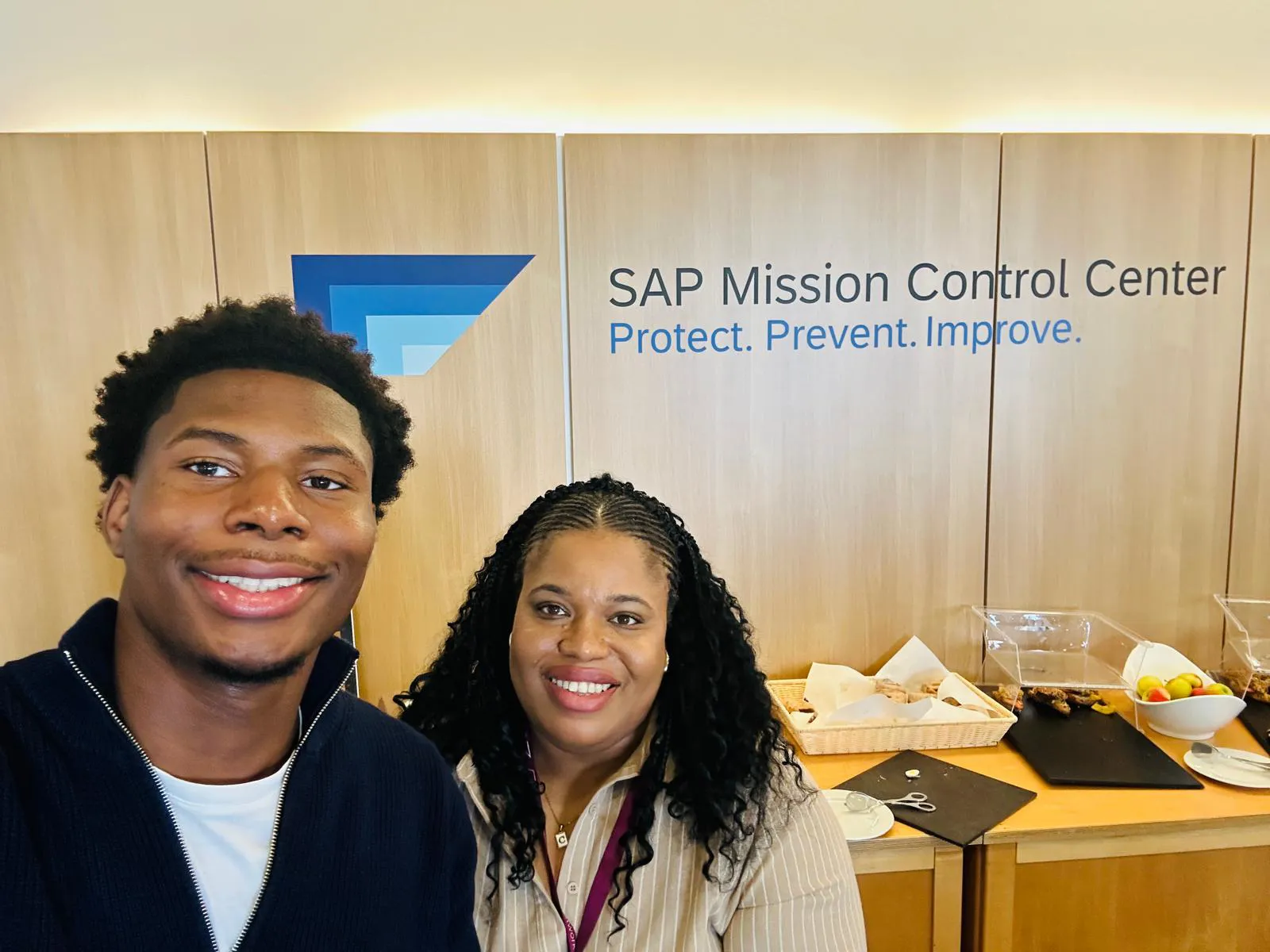 A young man and a woman pose for a selfie in front of a wall with the SAP Mission Control Center logo. The background includes text that reads "Protect. Prevent. Improve." and displays a table with snacks and fruit.