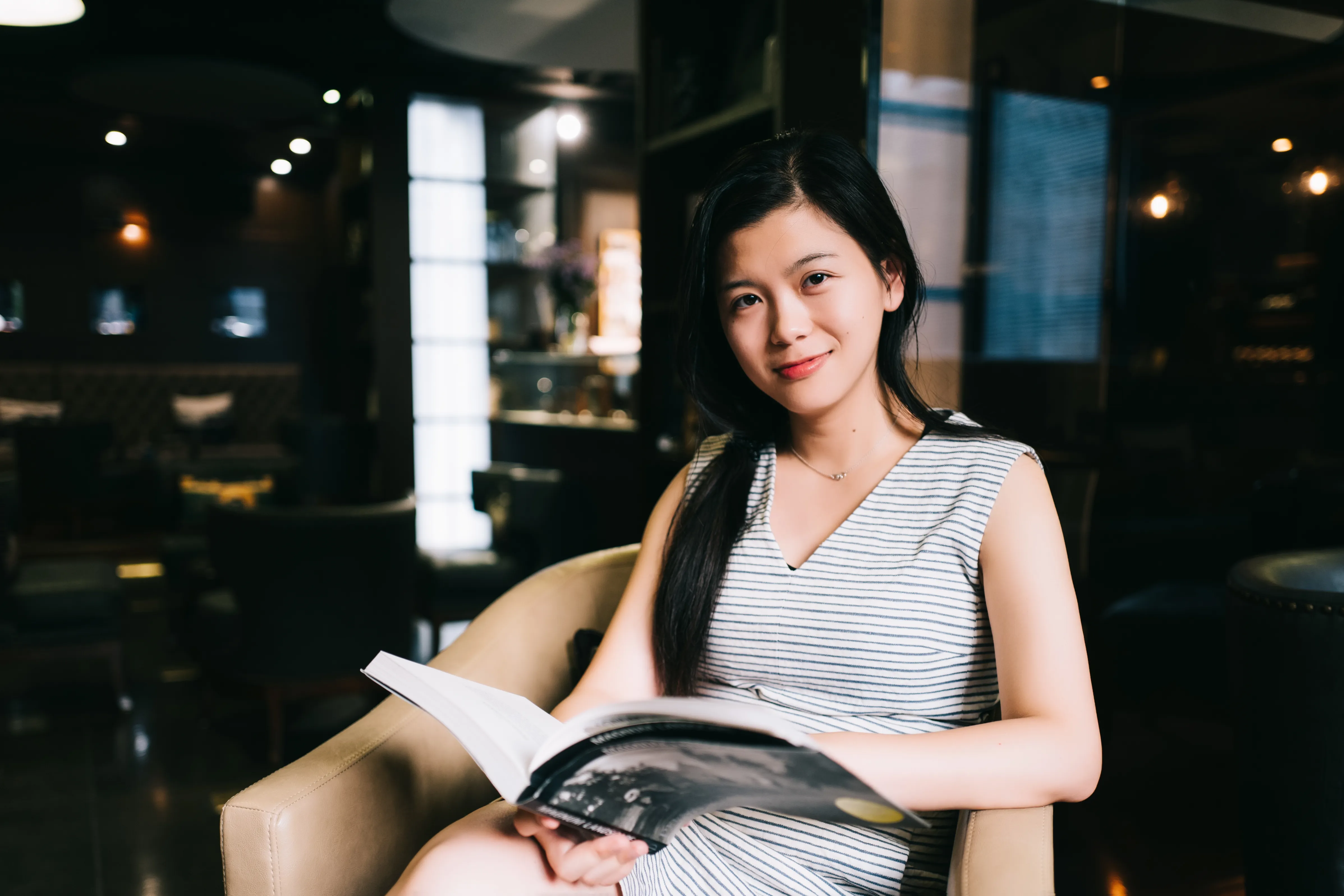 A woman in a striped dress is seated in a chair, holding an open book.