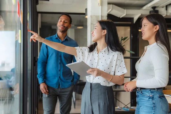Three colleagues discussing ideas in an office setting, with one pointing at sticky notes on a glass wall